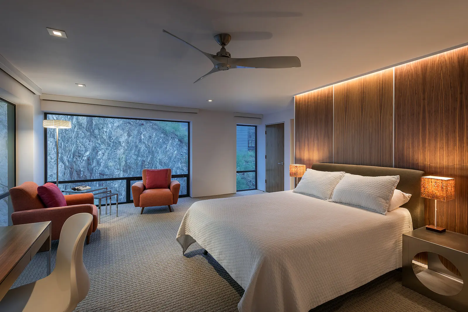 Modern bedroom with a large bed, wooden accent wall, and two orange chairs by a window.