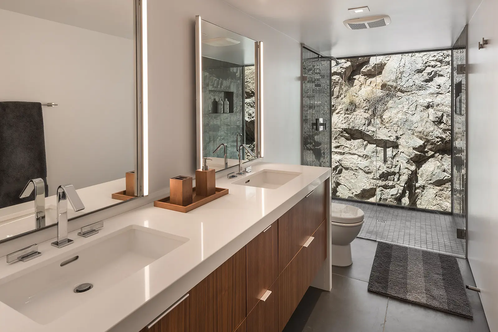 Modern bathroom with double sinks and glass-enclosed shower featuring stone wall.