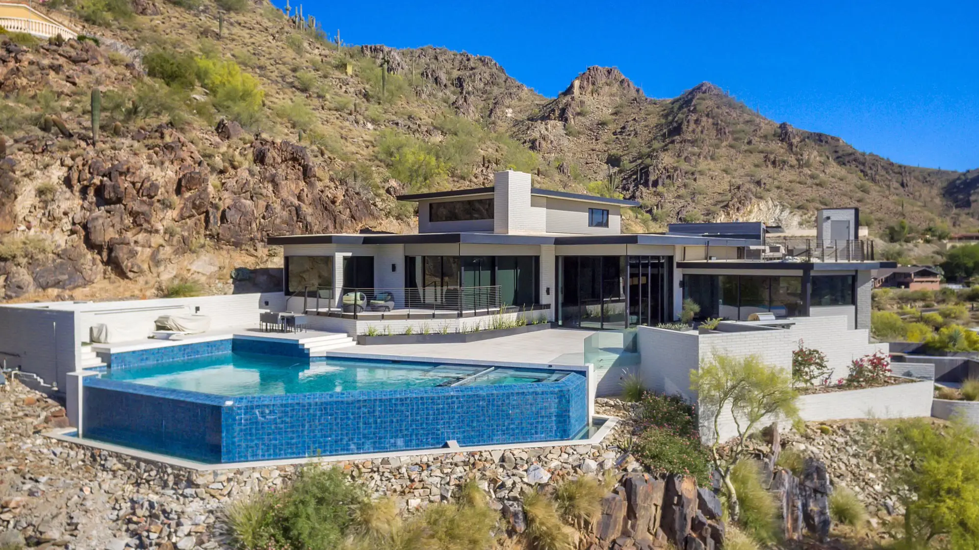 Modern house with an infinity pool overlooking rocky hills under a clear blue sky.